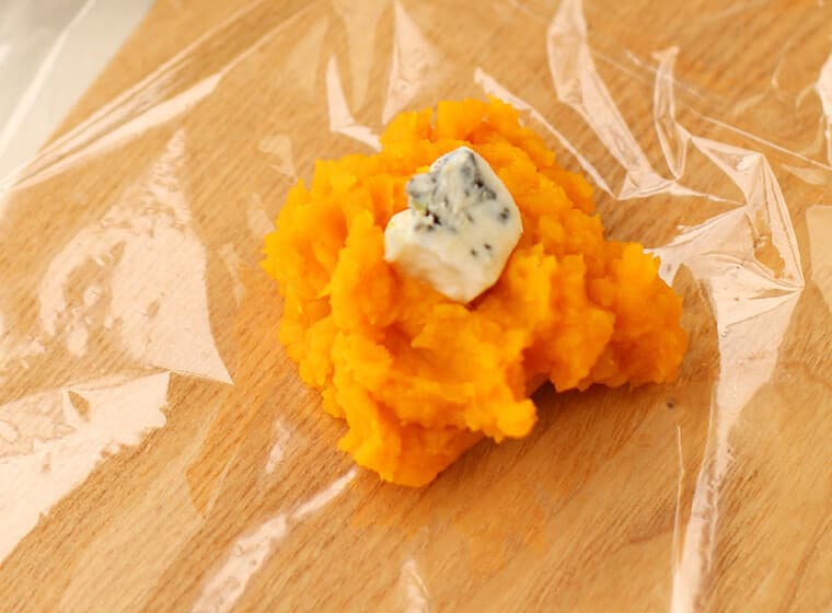 粗熱が取れたら6～8等分しラップの上に1個分ずつ広げ、中央にブルーチーズをのせ、ラップを絞って茶巾状にする（かぼちゃからチーズが少し見えるように包む）。